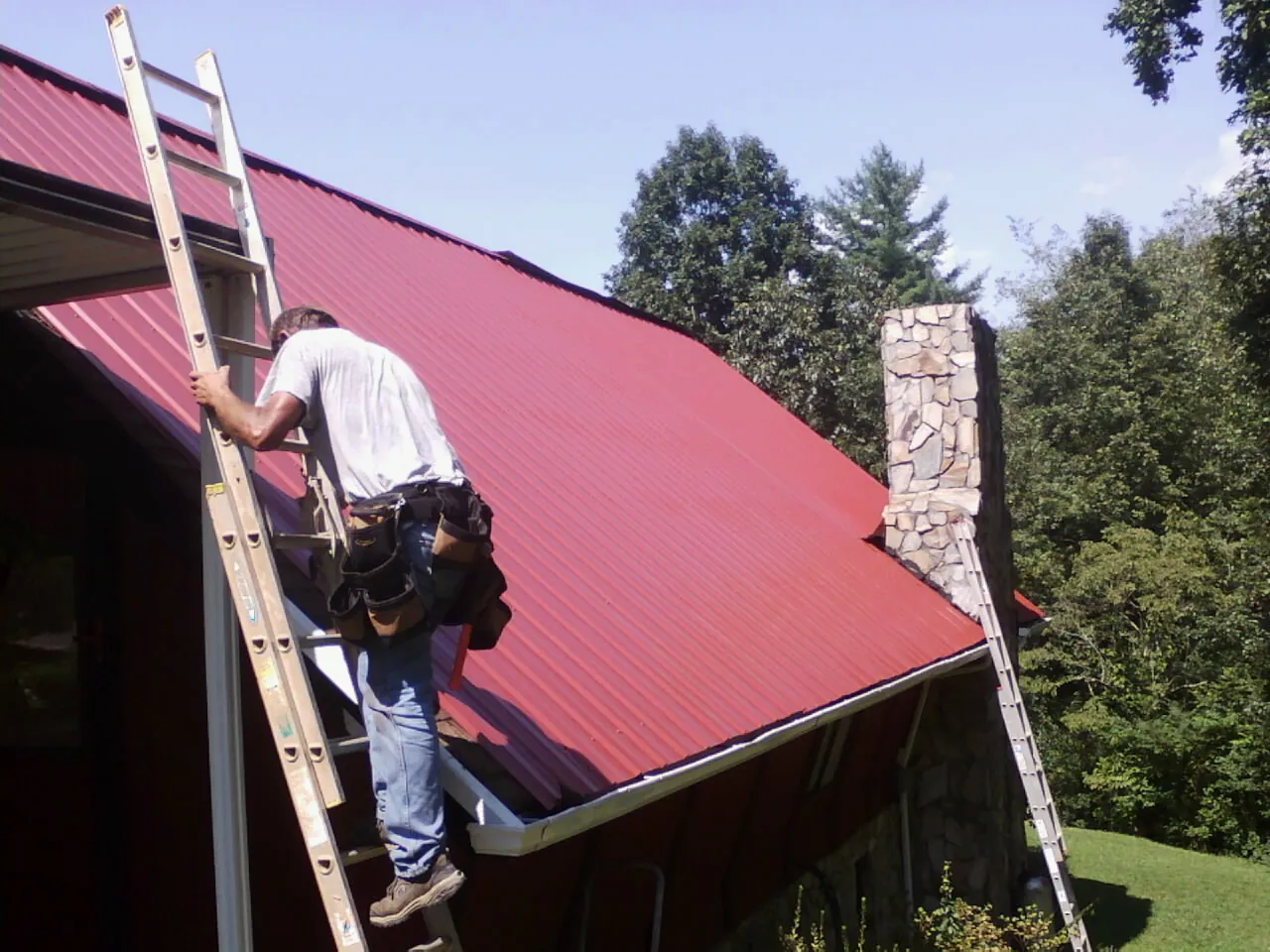 Roofer on a ladder beside a red metal roof and stone chimney — Soffit & Fascia Repair by Roofing Brothers in Montgomery Village